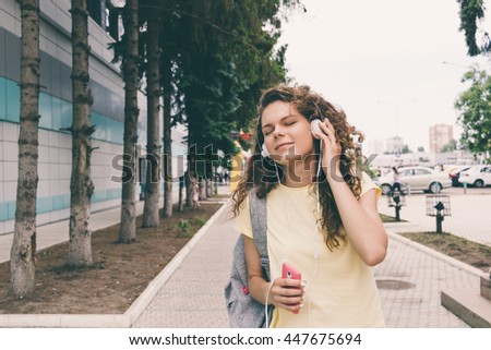 Curly brunette in a yellow T-shirt and headphones listening to music on the background of the city in summer. Happy woman outdoors.