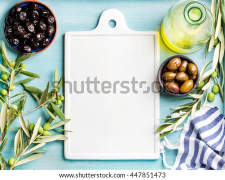 Two bowls with pickled green and black olives, olive tree sprigs and bottle of olive oil with white ceramic board in center over blue Turquoise background, copy space, top view