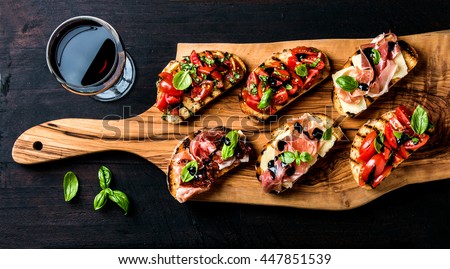 Brushetta set for wine. Variety of small sandwiches with prosciutto, tomatoes, parmesan cheese, fresh basil and balsamic creme served with glass of red wine on rustic wooden board over dark background Royalty-Free Stock Photo #447851539