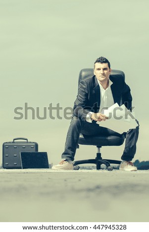 handsome male businessman in black formal jacket and white shirt with laptop and paper on leather arm chair outdoor with briefcase on cloudy sky background