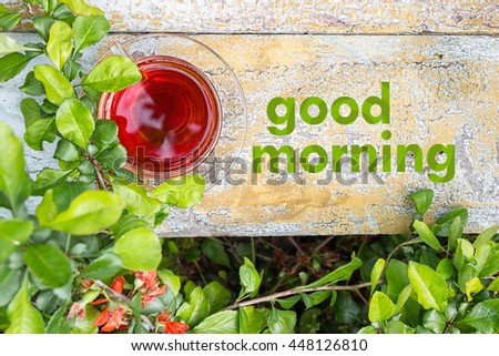 Glass cup of tea on a wooden surface, green grass around. Red orange flowers. Text Good morning on the background.