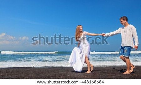Happy family on honeymoon holiday - just married young man and woman run with fun by black sand beach along sea surf. Active lifestyle, people outdoor activity on summer vacation on tropic island.