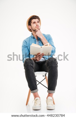 Thoughtful young man in hat sitting and reading a book over white background