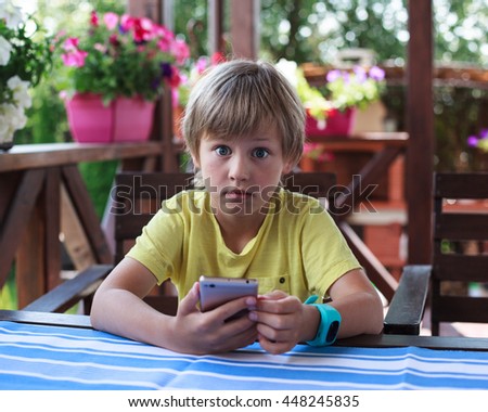 Cute boy with smart watch sitting in wooden armchair on a terrace and using mobile phone
