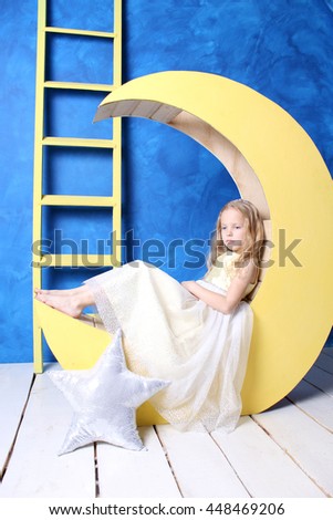 Sad blonde girl sitting on a yellow moon. The girl in a beautiful dress. 
Moon and ladder on a blue background. Girl dress with sequins