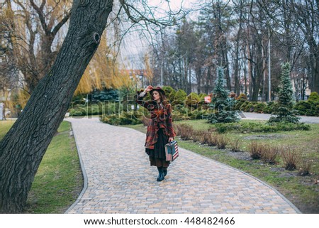 Beautiful young woman in a red hat and stylish dress with a suitcase with the American flag stands in the park on a background of trees and shrubs. English lady. Alone retro concept. 