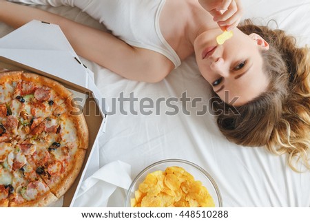 Girl eating chips on the bed, standing next to pizza Royalty-Free Stock Photo #448510288