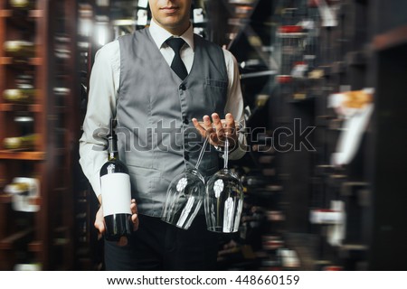 Close up of arms of skillful male sommelier standing in liquor store. He is holding a bottle of wine and two glasses. The man is smiling. Copy space in right side Royalty-Free Stock Photo #448660159