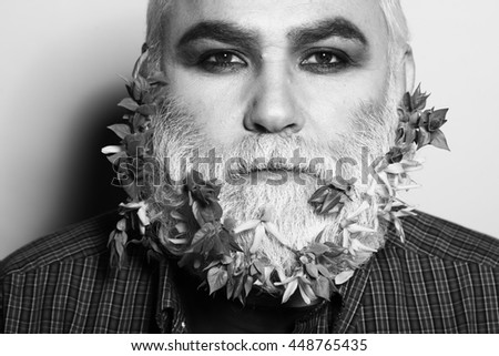 old bearded man with flowers and leaves in long grey beard on serious face in checkered shirt on studio background, black and white
