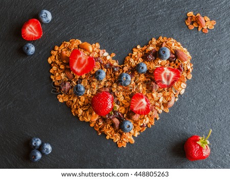 Ingredients in shape of heart to cook a breakfast. Blueberries, strawberries and granola made from oat flakes, dried fruits and nuts. Black stone background, top view.
