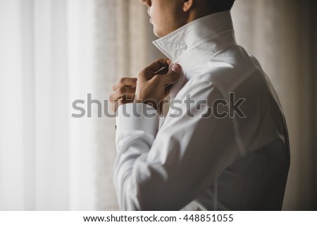 Man buttons up his white shirt standing in the front of a bright window Royalty-Free Stock Photo #448851055