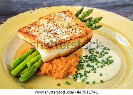 Halibut filet with asparagus sitting on serving of sweet potato puree and lemon butter sauce garnished with chives on rustic yellow plate