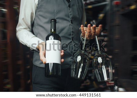 Close up of arms of skillful male sommelier standing in liquor store. He is holding a bottle of wine and two glasses. The man is smiling. Copy space in right side Royalty-Free Stock Photo #449131561