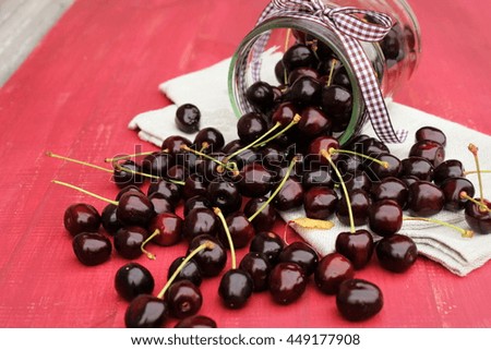 Glass jar full of ripe tasty black cherry with grey linen cloth on red wooden table. Composition with berries in bright color with rustic decor elements