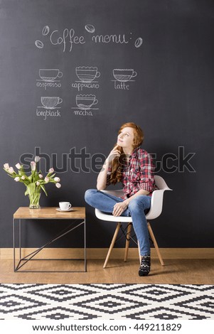Ginger woman sitting beside new small table, in the background blackboard coffee menu
