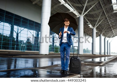 Picture of  young  businessman holding  suitcase and umbrella looking on watch waiting at rainy station