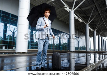 Picture of  confident young redhaired businessman holding black umbrella and suitcase in rain at terminal