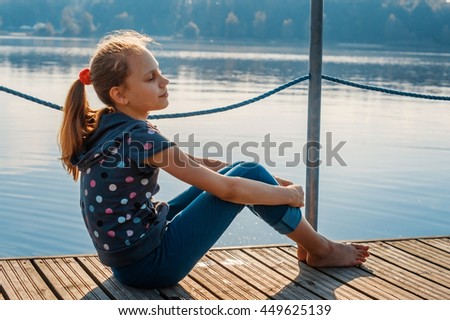 Child girl sitting on a wooden bridge near the water on the lake. At sunset in the evening