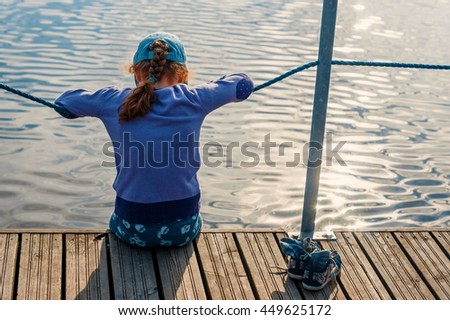 Child girl sitting on a wooden bridge near the water on the lake. At sunset in the evening