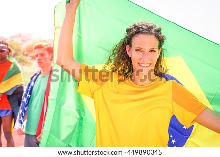 Brazilian woman sport fan holding flag at stadium  with american and jamaican athletes background -  Cheerful supporter girl with blonde curly hair looking camera - Concept of competition victory