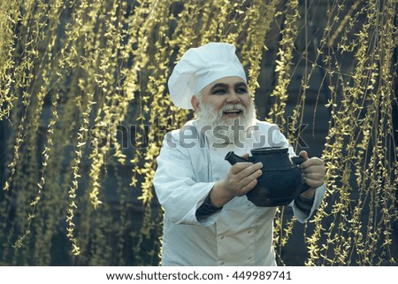 bearded man cook chef in uniform and hat with long beard on smiling face holding iron old tea kettle in blooming tree branches outdoor on natural background