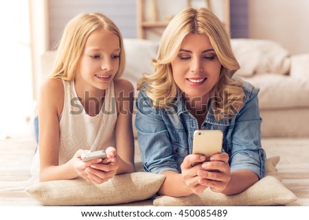 Portrait of beautiful blonde woman in jeans shirt and her teenage daughter using phones and smiling while lying on the floor at home