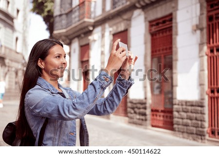 Sweet young girl taking pictures in a narrow street with a cell phone wearing her hair loose and casual clothing