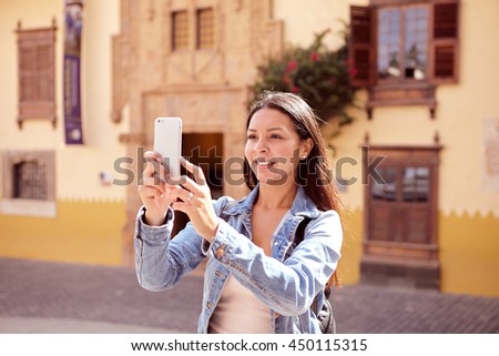 Pretty young girl standing, taking a picture with a cellphone while smiling and her hair loose while wearing a denim jacket