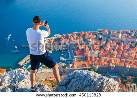 Male traveller make a panoramic photo of the old city of Dubrovnik from the mountain to his phone for Instagram or other social networks. Travel to Croatia. Summer vacation.