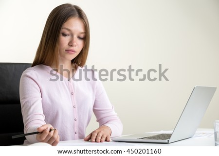 Young woman using laptop and doing paperwork on office desktop. Concrete wall background