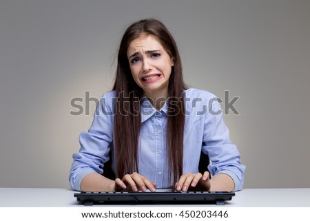 Playful young woman with funny regretful expression using computer keyboard on grey office desktop