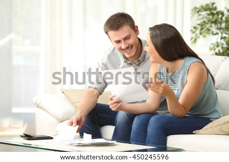 Happy couple reading mail and checking accountancy looking each other sitting on a couch at home Royalty-Free Stock Photo #450245596