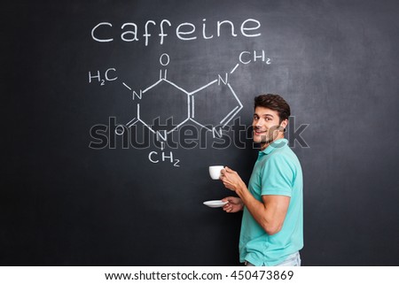 Happy young scientist standing over chemical structure of caffeine molecule drawn on chalkboard background and drinking coffee