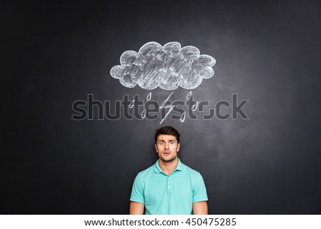 Surprised astonished young man standing under raincloud with lightning and rain drawn on blackboard background