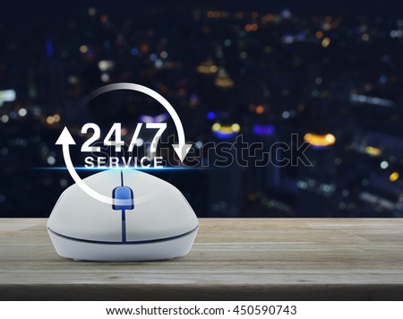 Wireless computer mouse with button 24 hours service icon on wooden table in front of blurred light city tower, Full time service concept