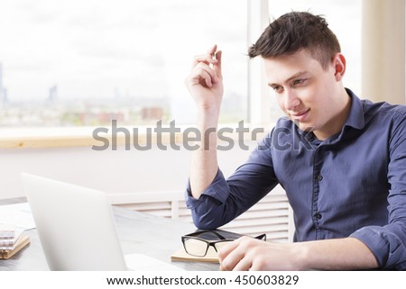 Young man with pen in hand looking at laptop screen placed on wooden desktop with glasses and notepad with window and blurry city view in the background