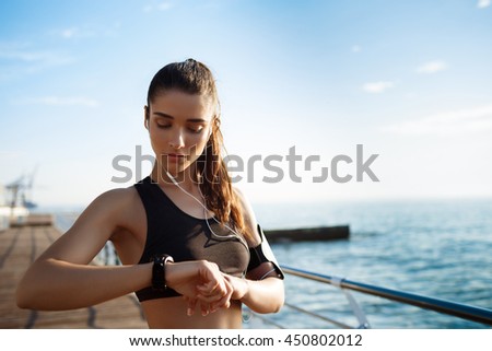 Picture of young attractive fitness girl looking at watch with sea coast on background