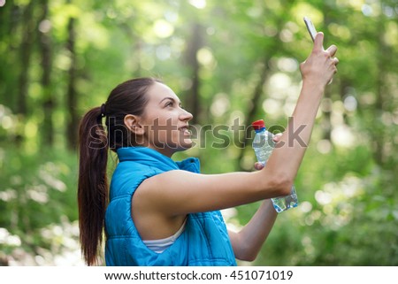 Smiling beautiful young running sporty woman macking selfie with a bottle of water on the nature, forest, park background in blue sleeveless jacket.Sport. Instagrram