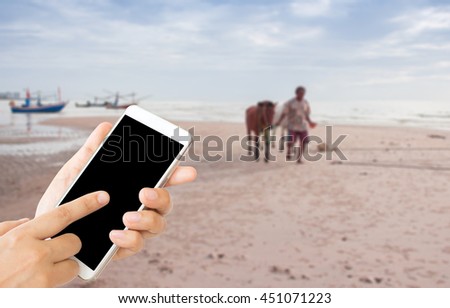 woman use mobile phone and blurred image of little old man lead horses on the beach in cloudy morning