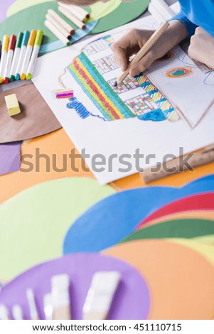 Close-up of kid coloring picture of boat. Next to him on a desk colorful sheets of paper and pens