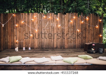 Wooden wall decorated by electric lamps and Love sign. Vintage suitcases and pillows on floor. Wedding. Reception. Lounge zone. Royalty-Free Stock Photo #451137268