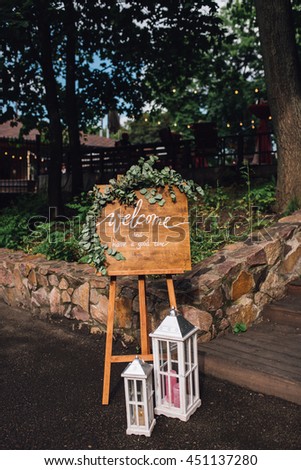 Wedding. Reception. Handmade wooden board with welcome sign on it decorated with eucalyptus on wooden stand with lanterns on side.
