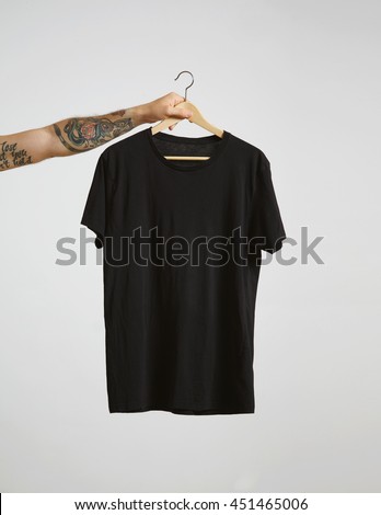 Tattooed biker hand holds hang with blank black t-shirt from premium thin cotton, isolated on white mockup Royalty-Free Stock Photo #451465006