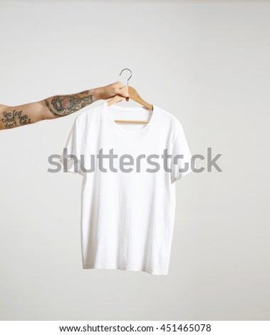 Tattooed biker hand holds hang with blank white t-shirt from premium thin cotton, isolated on white mockup Royalty-Free Stock Photo #451465078