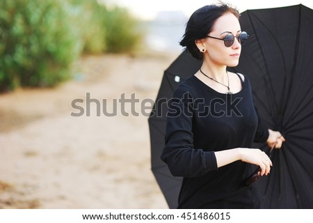 Beautiful young brunette girl in black clothes with a large black umbrella on the shore of the Bay the cool summer day.