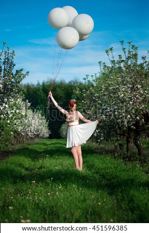 A picture of a young girl in a green garden holding huge balloons up high and waving her white skirt smiling down