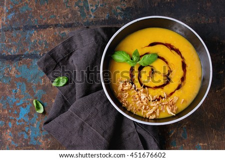 Black ceramic bowl of pumpkin and sweet potato cream soup with fresh basil, fried onion and balsamic sauce, served on textile napkin over old wood textured background. Top view