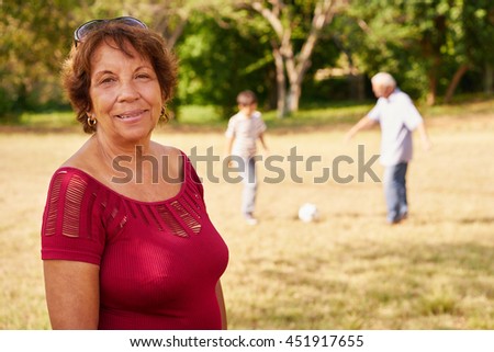 Portrait of confident and happy grandmother, smiling at camera while her grandson and husband play football in background. Concept of grandparents spending time with boys.