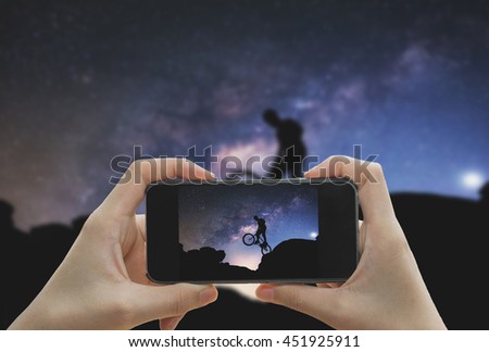 Taking photo on smart phone concept.Man riding a bike performing a trick against on Mountain with Milky way. 