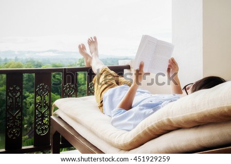 Rear view of asian man relaxing on a sofa and holding book on bed at home terrace with beautiful green background view. Relaxing concept.

